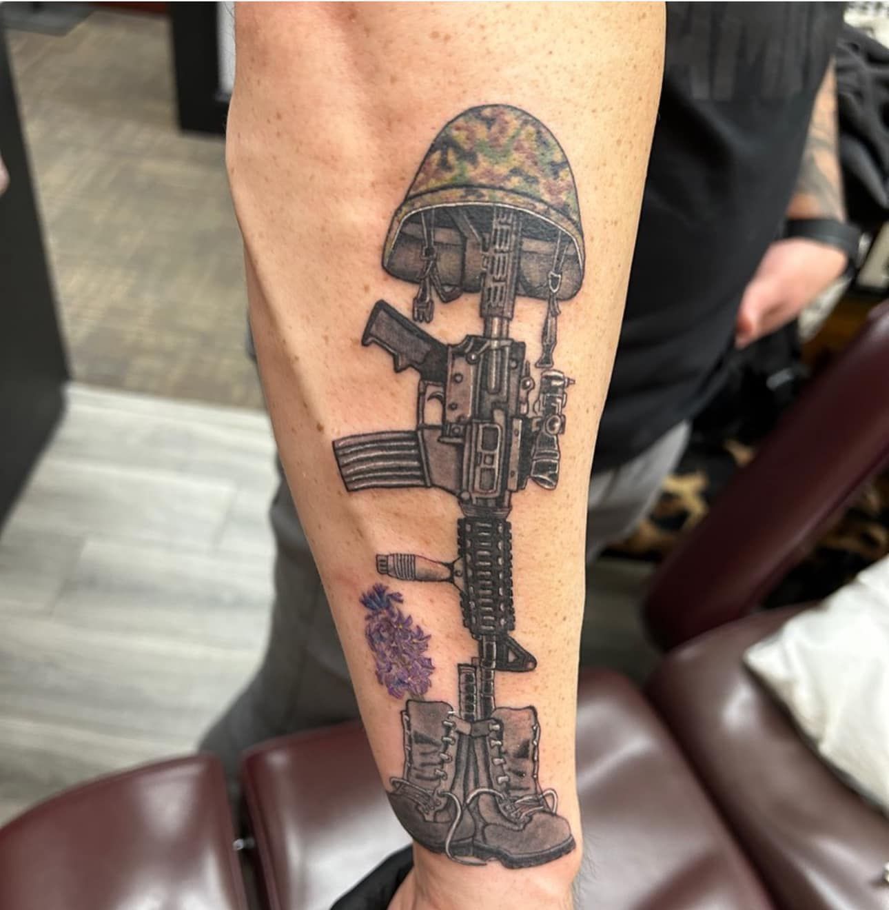 Forearm tattoo of a rifle assembled vertically with camo helmet, boots, and purple flowers