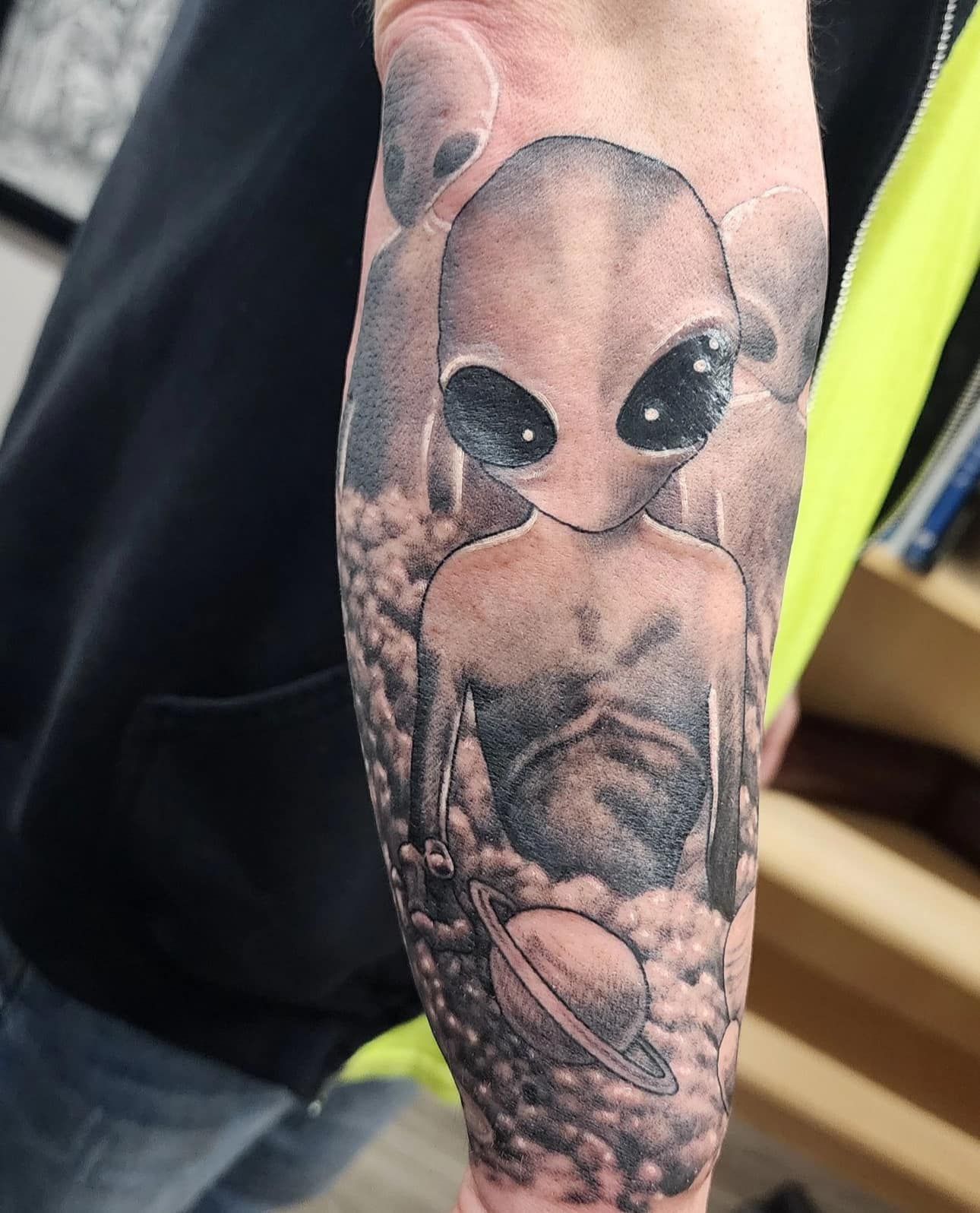 Tattoo of a gray alien with large black eyes and a small body on a forearm