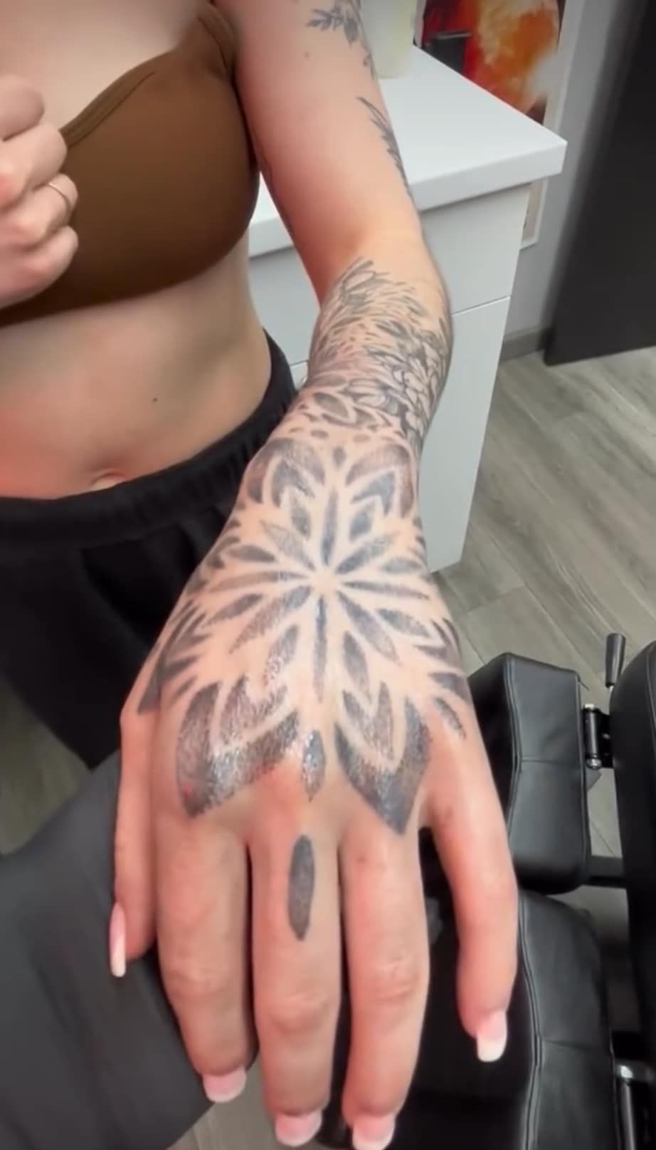 Hand with black-and-gray floral tattoo resting on a salon chair arm, with manicured nails visible
