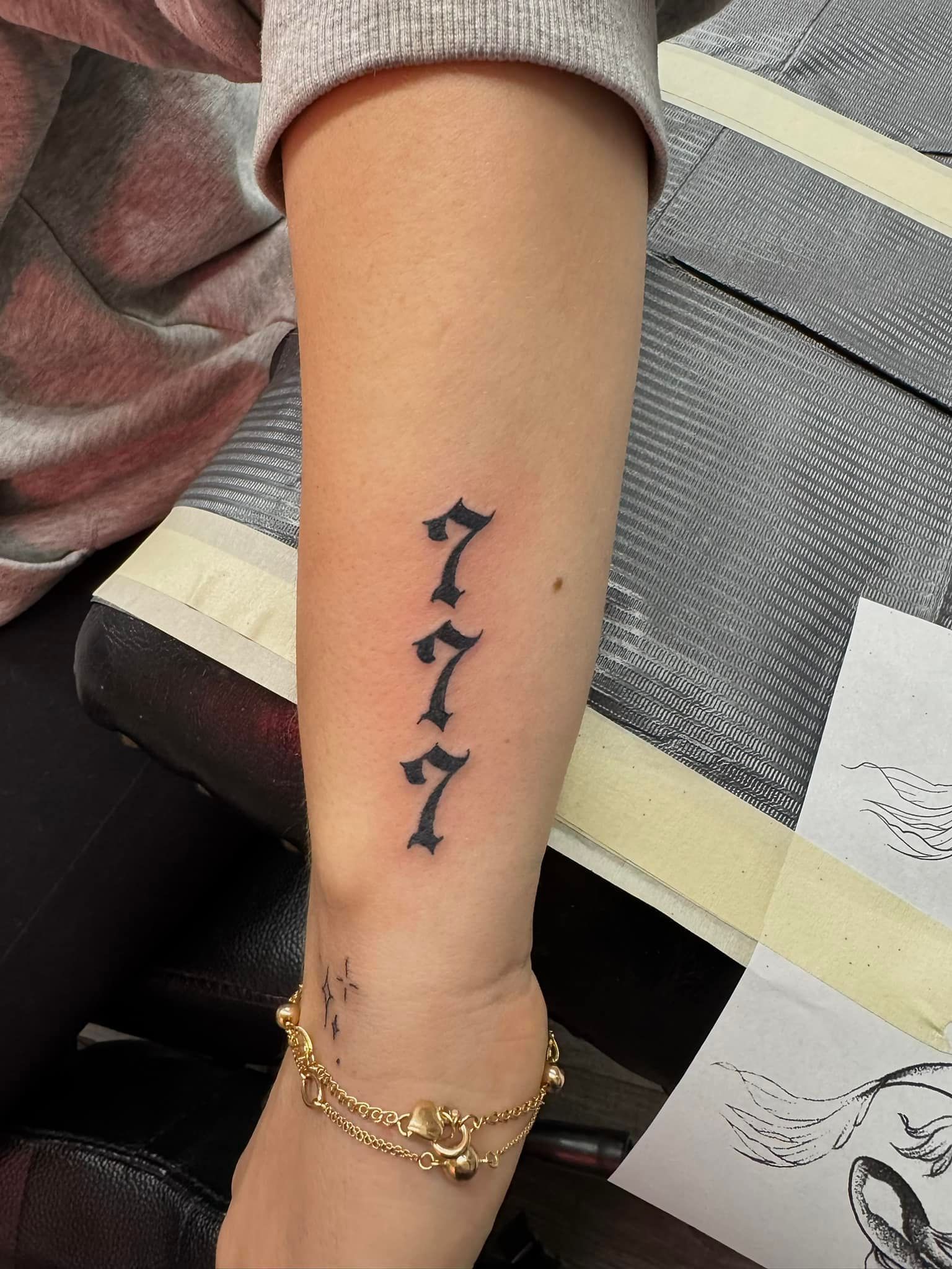 Close-up of a forearm with a vertical black tattoo of three sevens and a gold bracelet.
