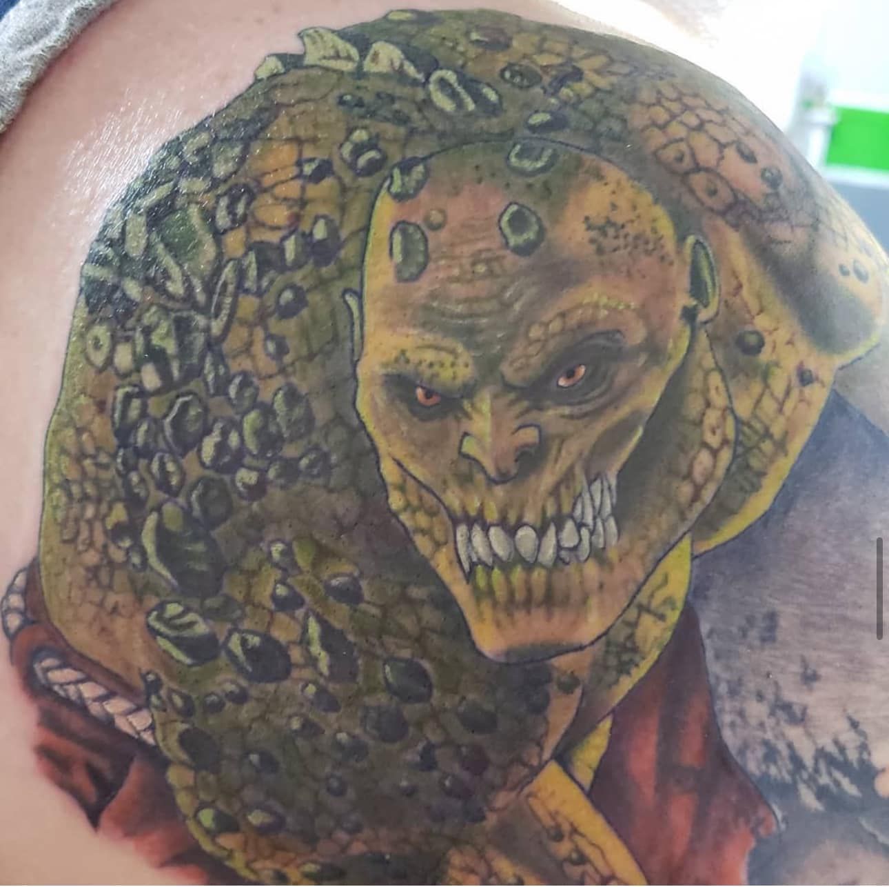 Detailed tattoo of a menacing skull-like creature with yellow eyes and scaly green skin on a shoulder