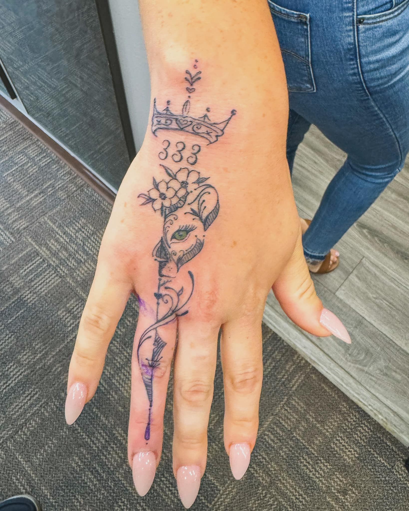 Close-up of a hand with a black floral tattoo and long pink nails on a carpeted floor