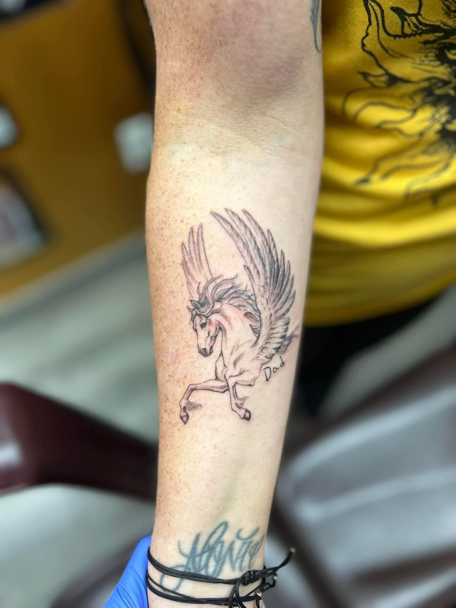 Close-up of a forearm with a small black line-art tattoo of a winged figure, resting on a chair