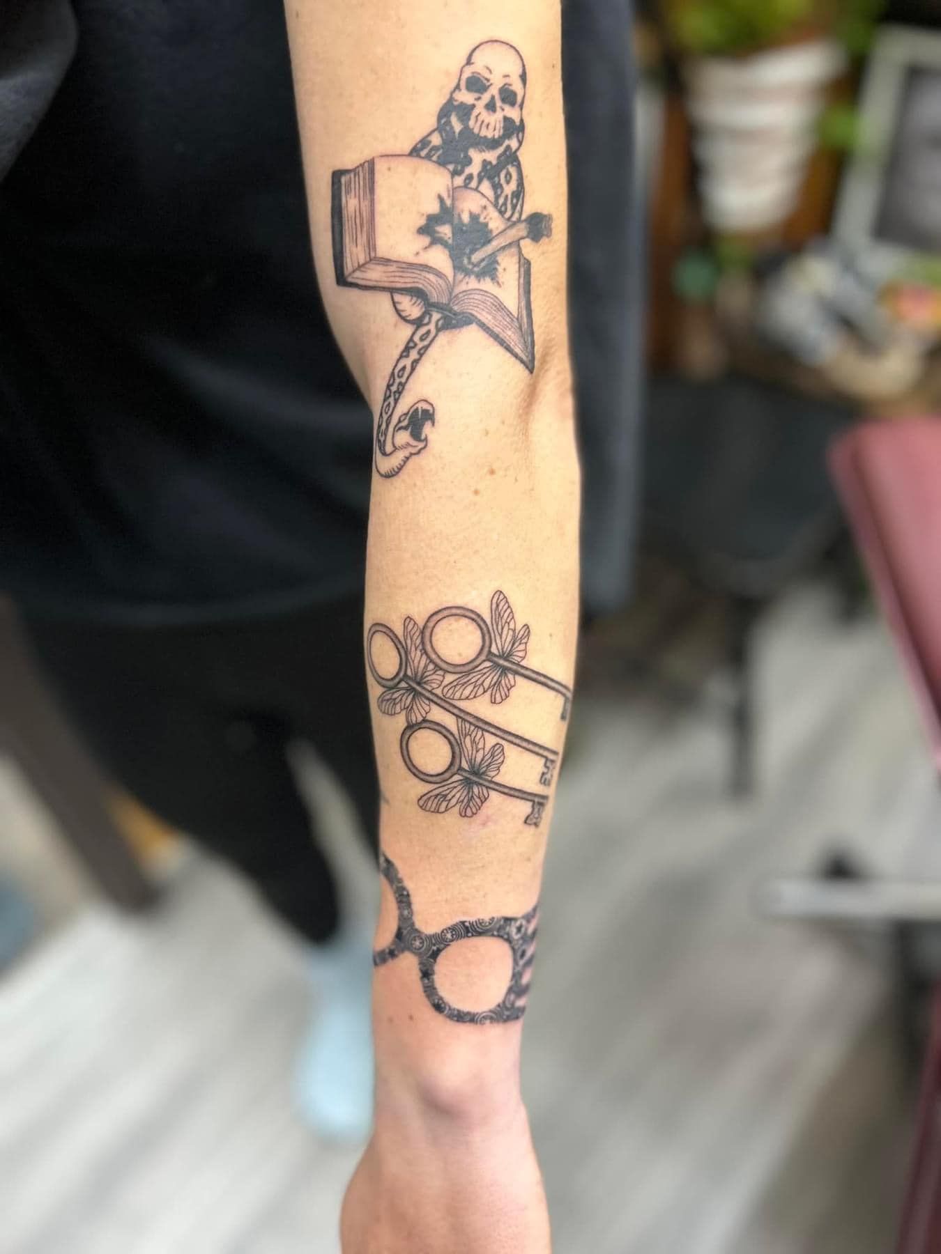 Forearm with dark linework tattoos, including a skull, gears, and a circular symbol on a blurred indoor background
