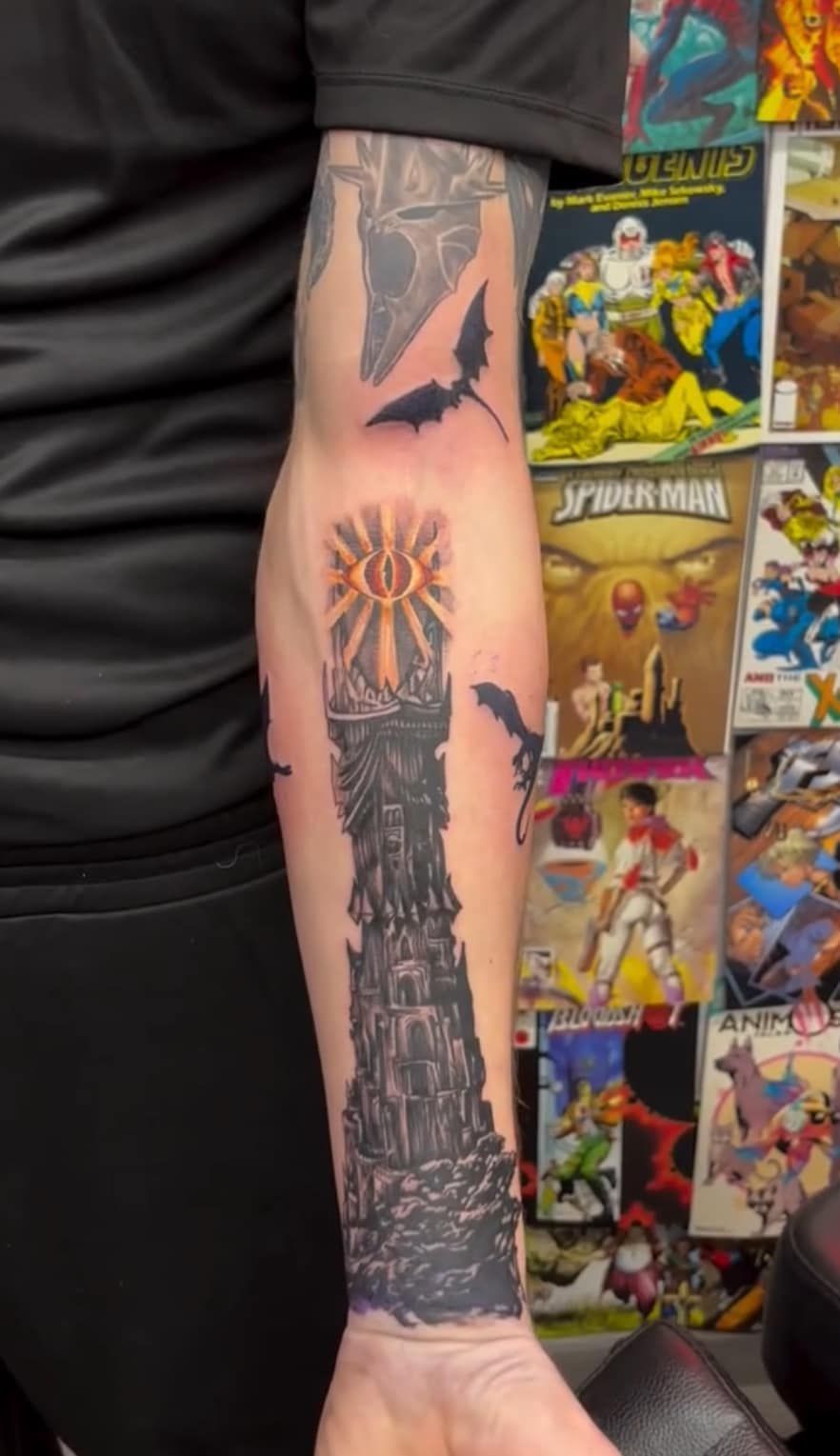 Close-up of a tattooed forearm with a black and orange sleeve tattoo beside a colorful poster wall