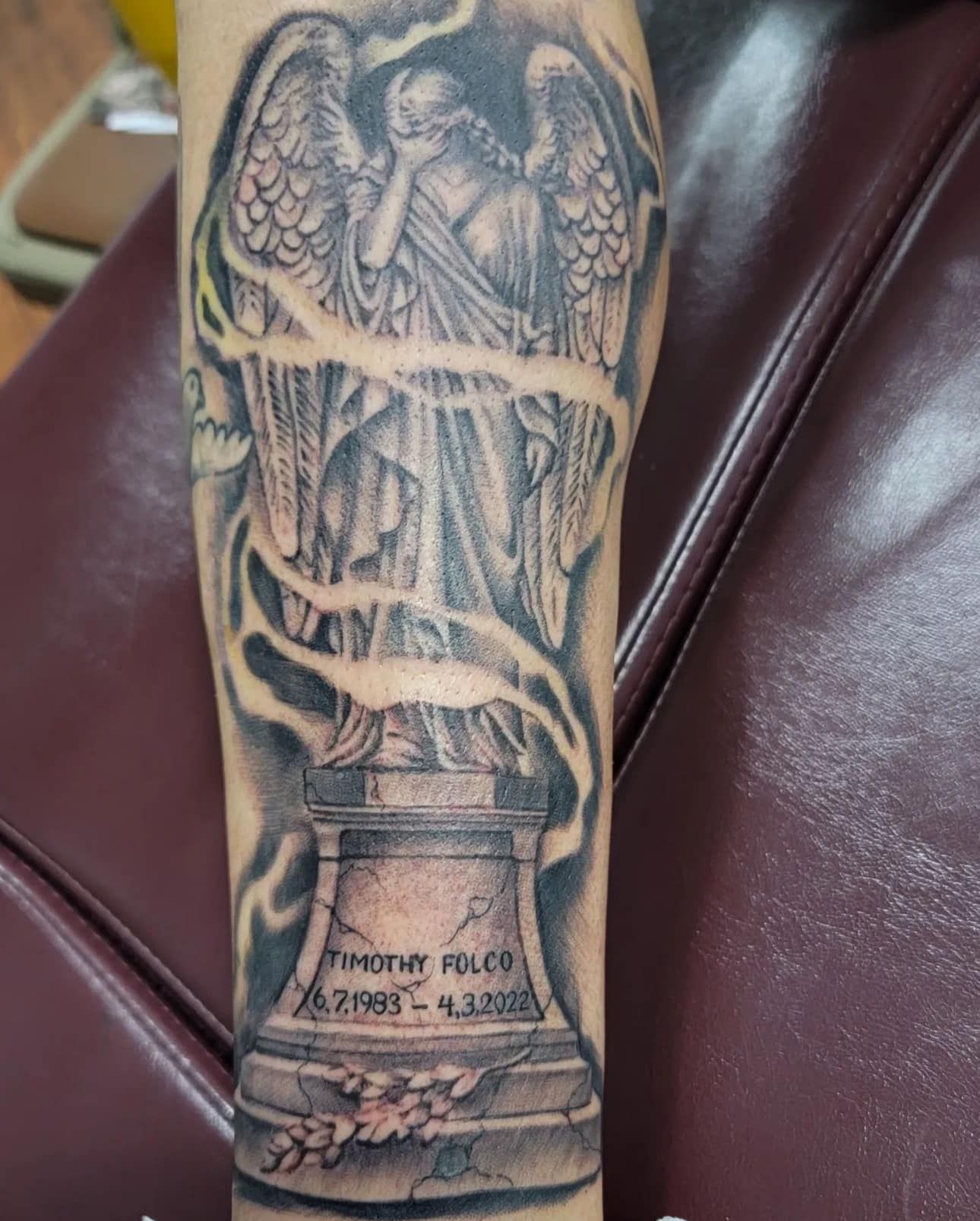 Black-and-gray angel tattoo on a forearm, with flowing fabric and pedestal details.
