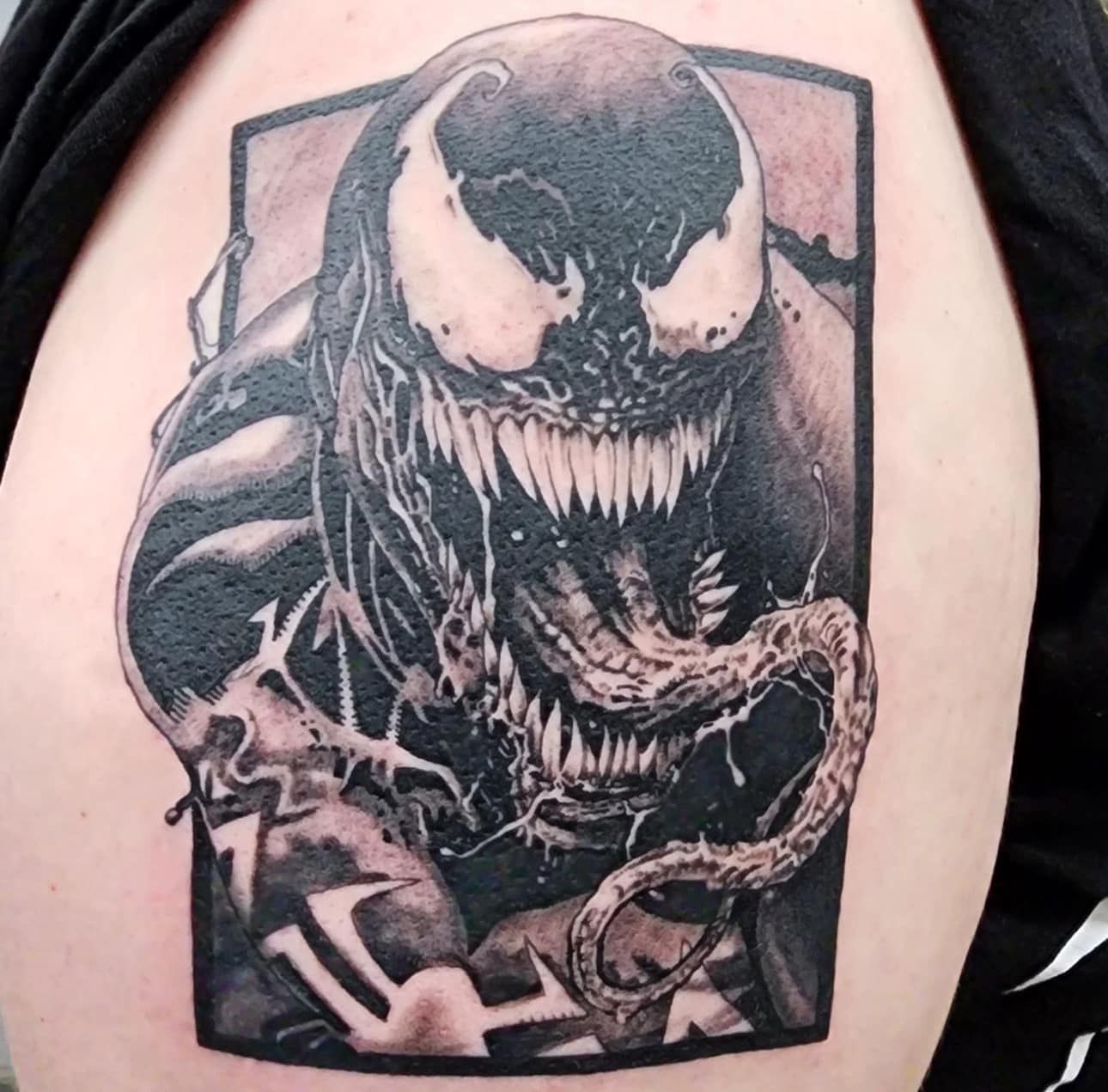 Black-and-gray Venom tattoo on an upper arm, showing a snarling symbiote face.