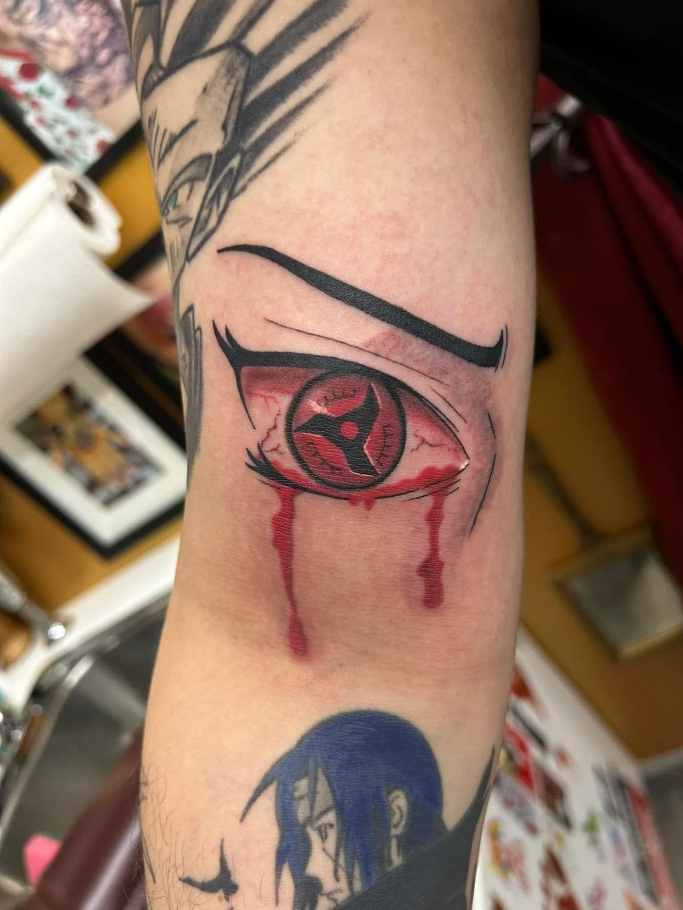 Close-up of a forearm tattoo of a red eye with black lines and red drips, above a blue tattoo below