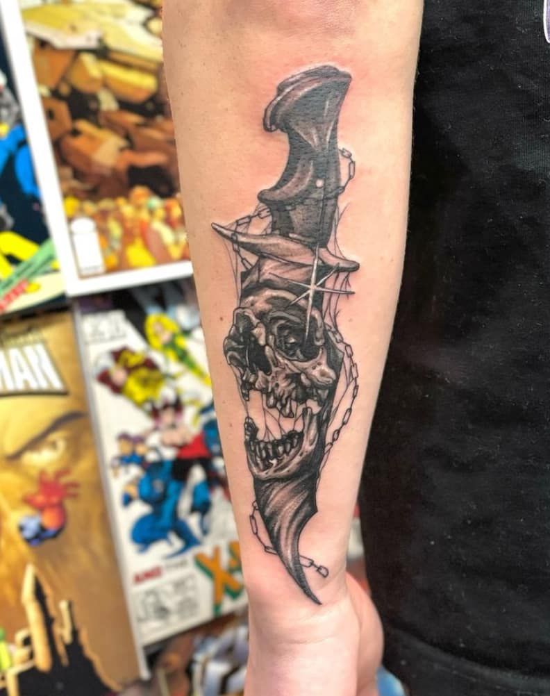 Arm tattoo of a detailed black-and-gray dagger with skull-like face on a forearm