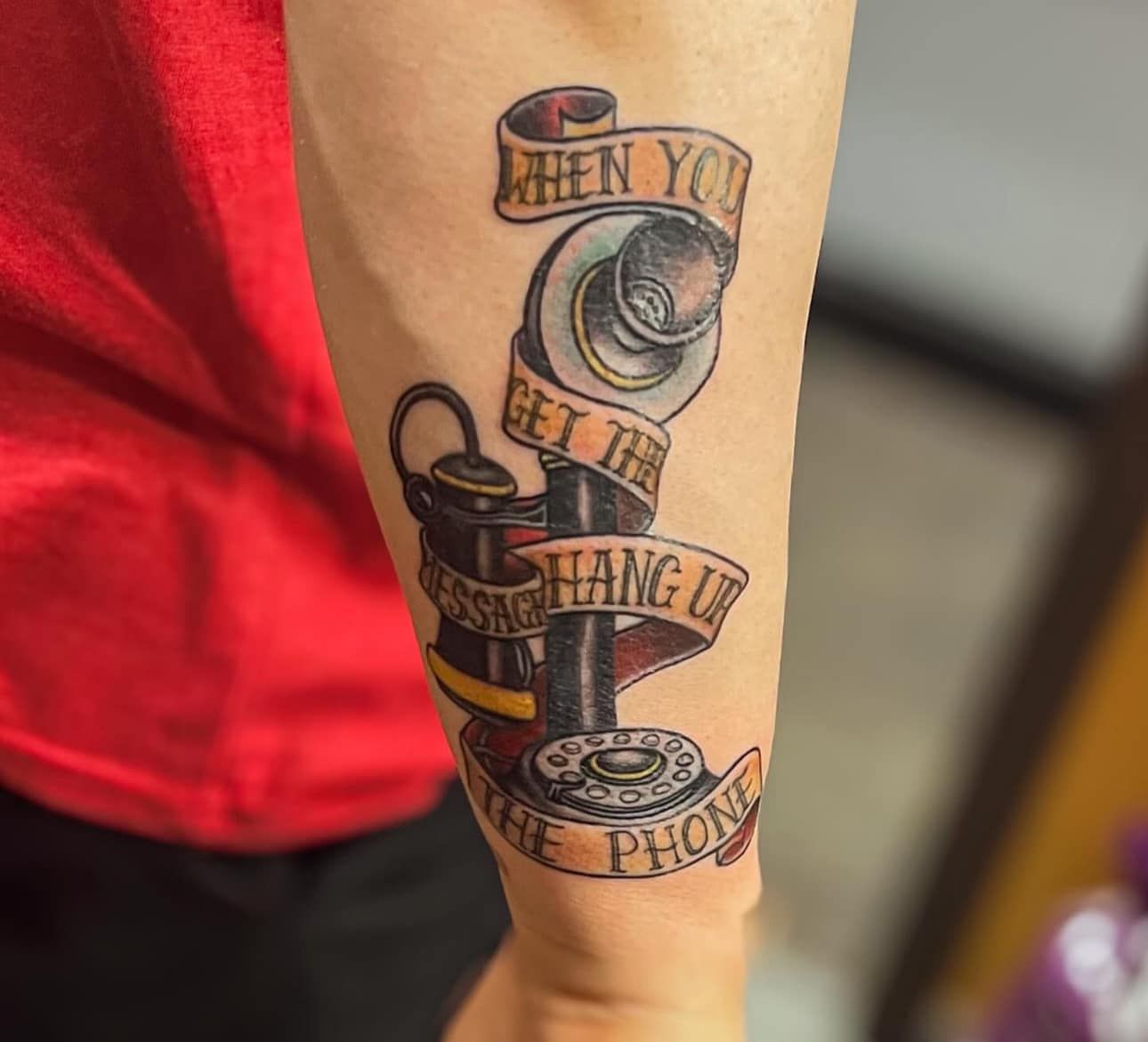 Forearm tattoo of a vintage camera with ribbon text, on a person wearing a red shirt