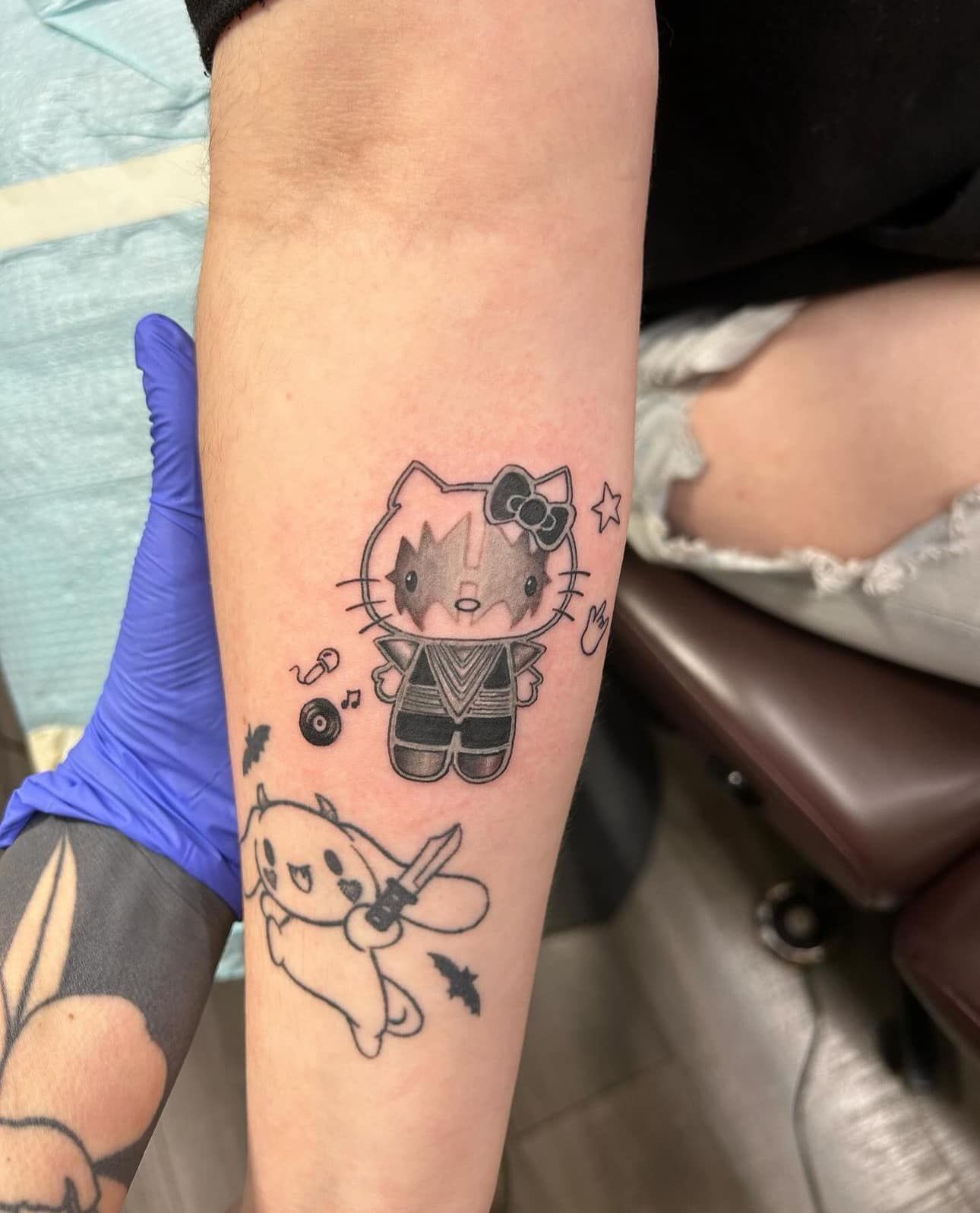 Tattooed arm with blue-gloved hand, showing a Hello Kitty tattoo and a bunny outline tattoo