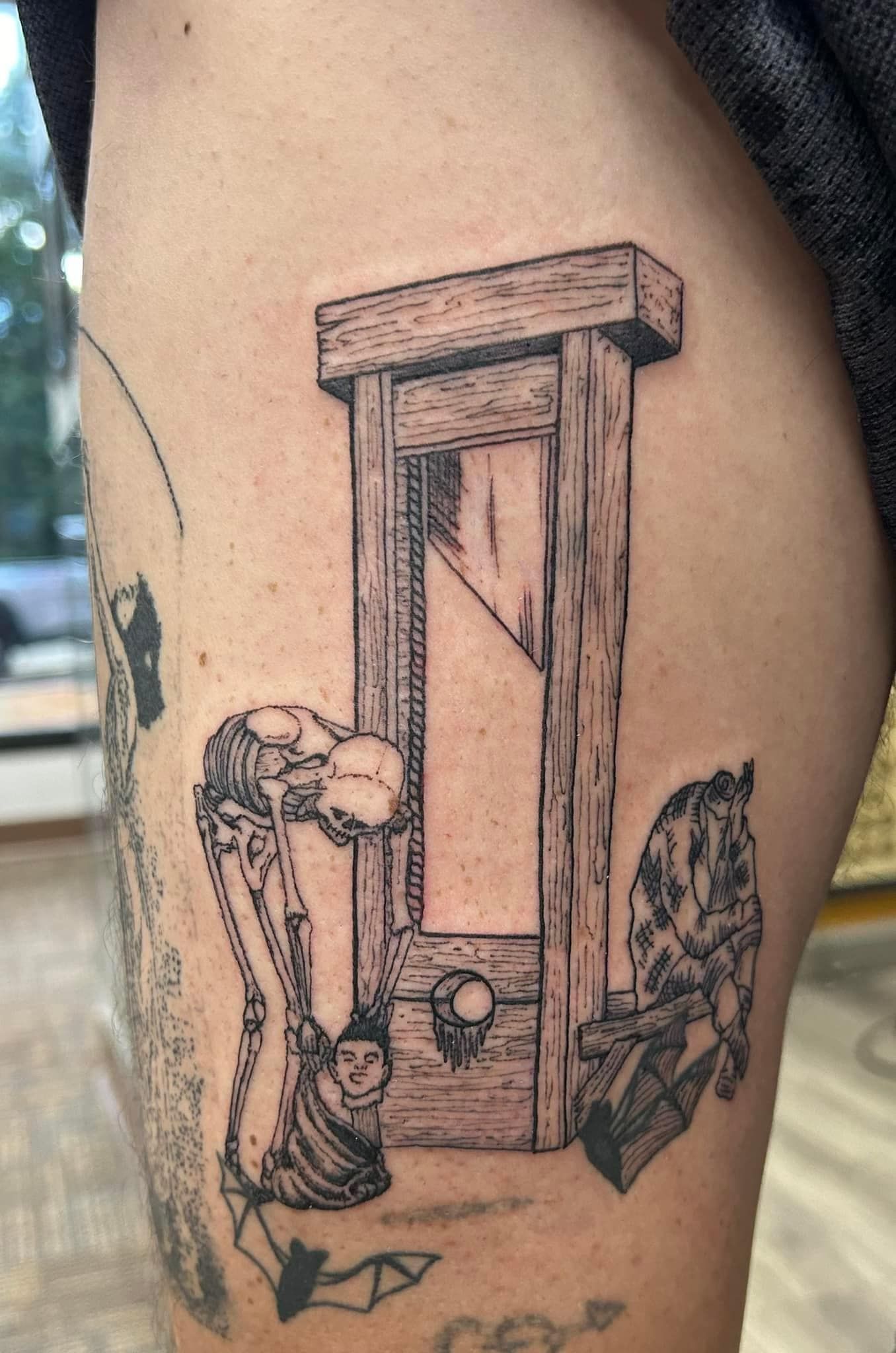 Black linework tattoo of a guillotine with two figures on a person’s arm
