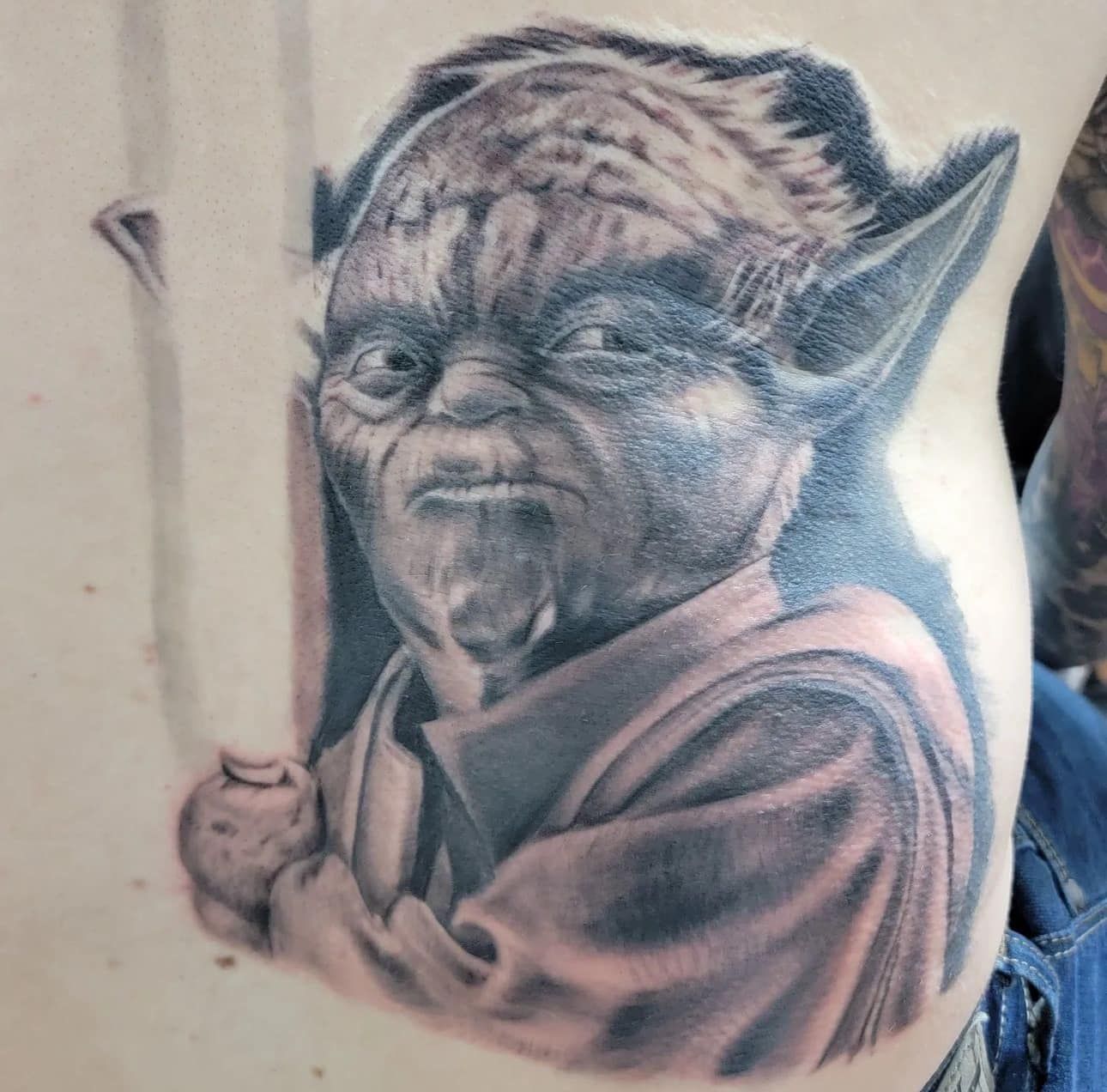 Grayscale tattoo of Yoda holding a light saber, showing a stern expression.