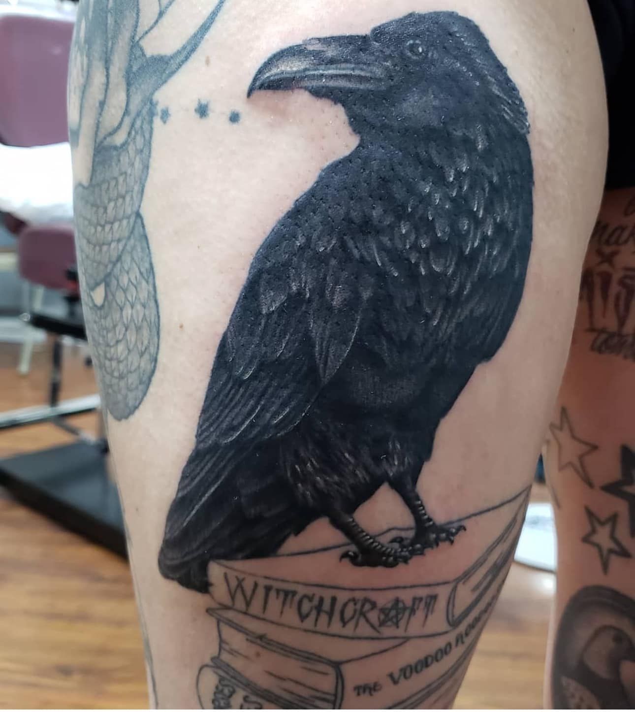 Black raven tattoo on a person’s upper arm above a banner reading “Witchcraft”