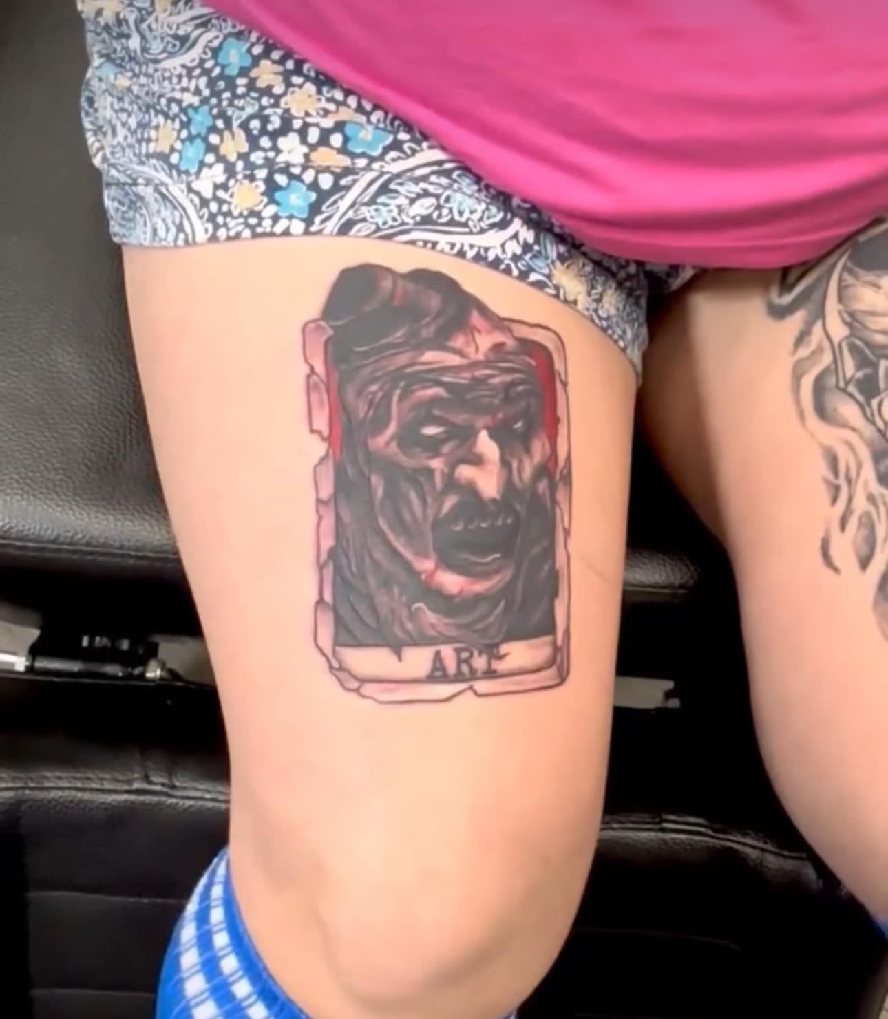 Thigh tattoo of a snarling dog in a shaded rectangular frame, with blue patterned shorts and pink shirt visible