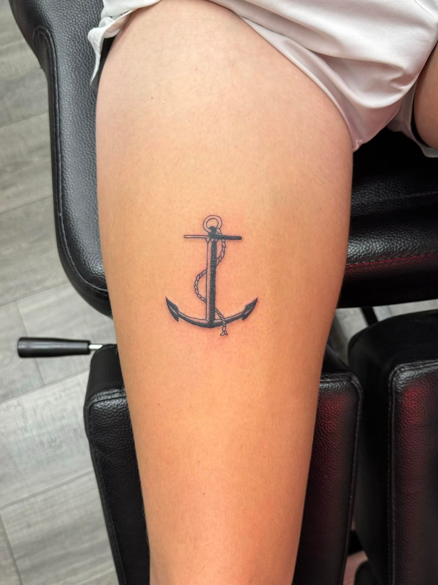 Close-up of a small black anchor tattoo on a person’s upper thigh, beside a chair.