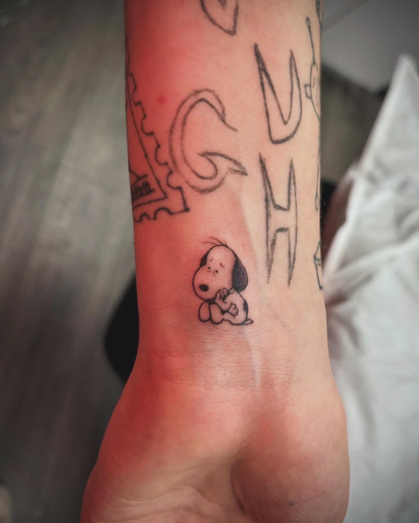 Close-up of a wrist tattoo featuring a small Snoopy-like cartoon dog and a large outlined letter design.