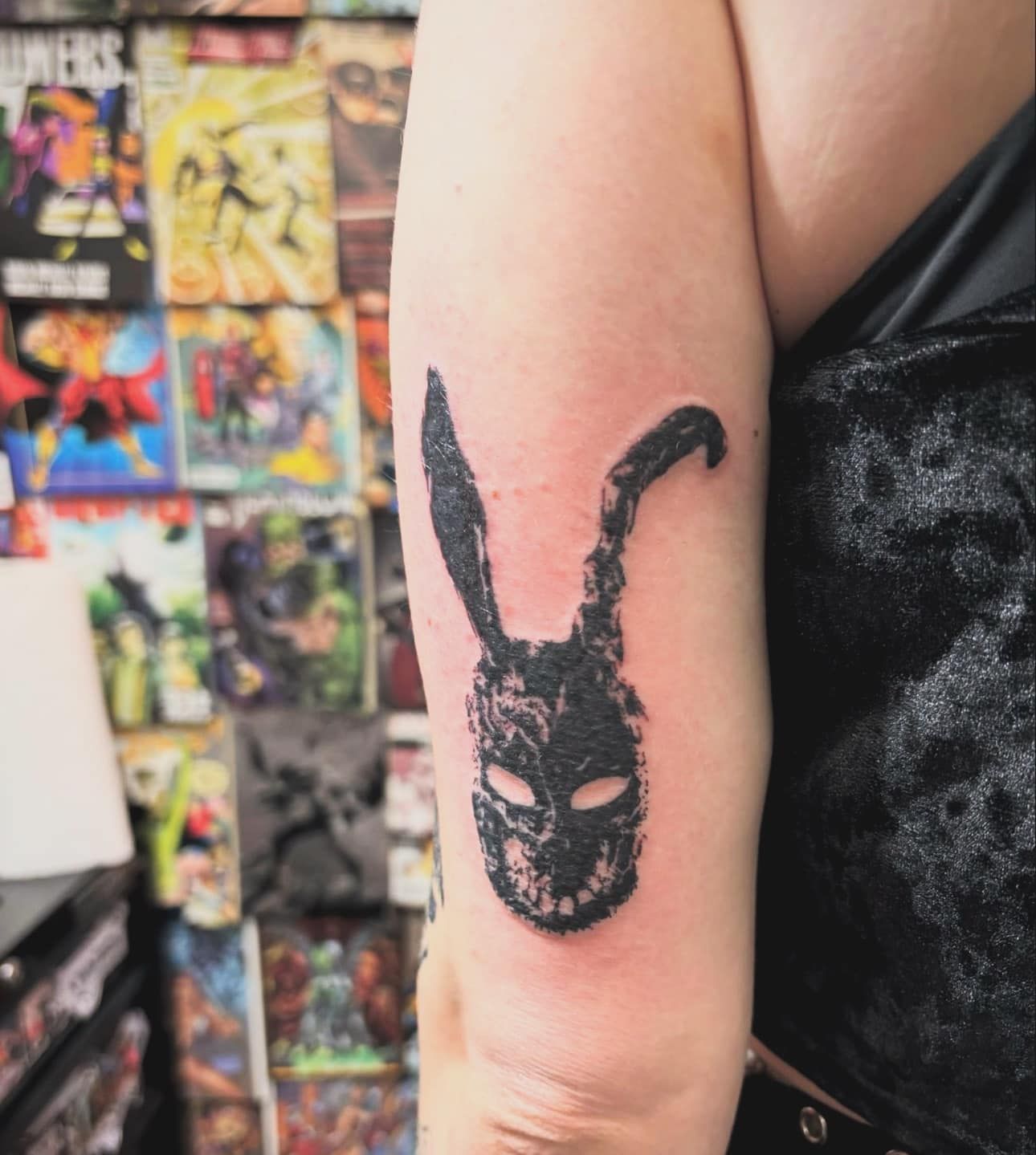 Close-up of an upper arm with a black tattoo of a bunny-faced mask, against a colorful poster wall