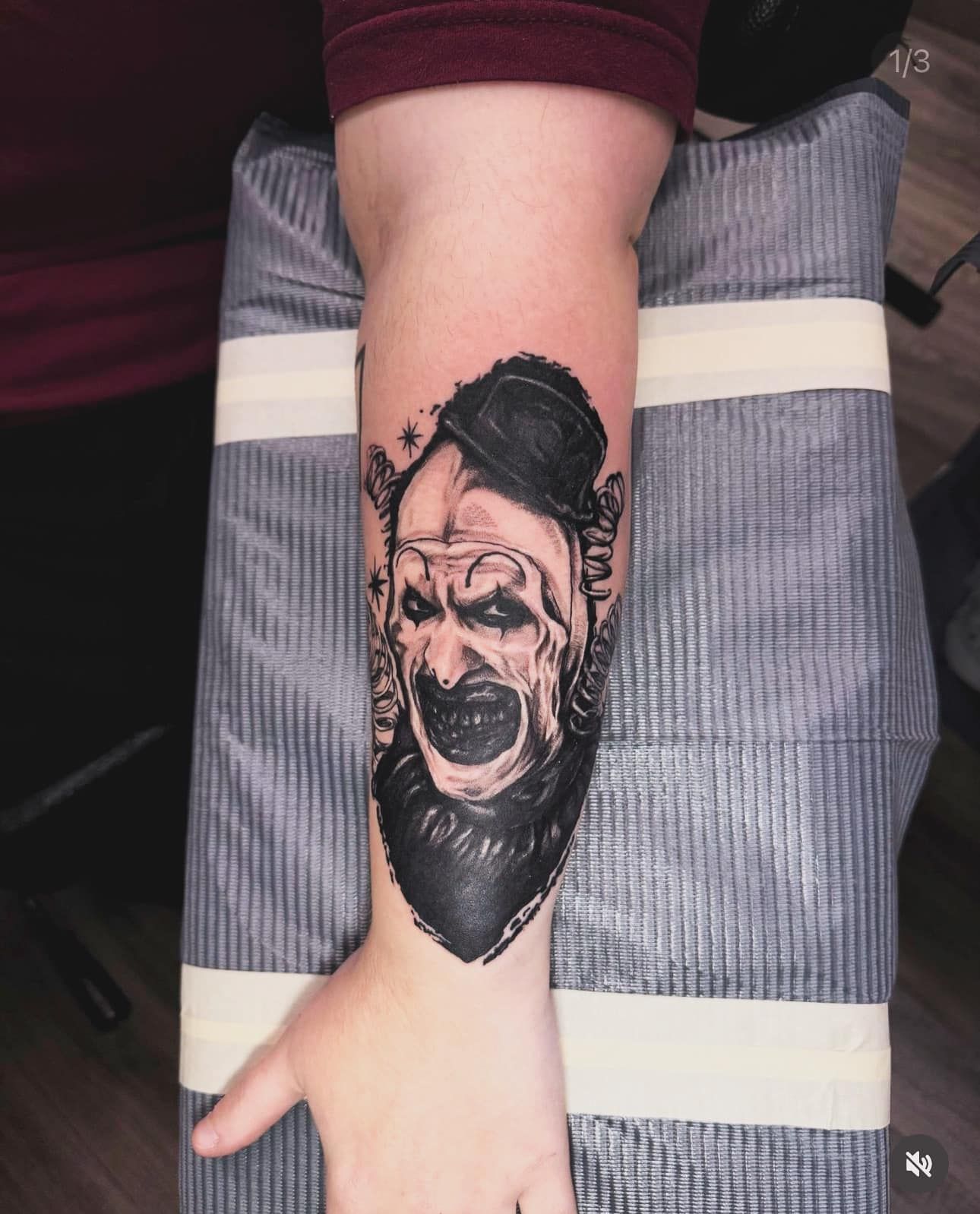 Arm tattoo of a bearded face in black ink on a forearm resting on a gray chair.