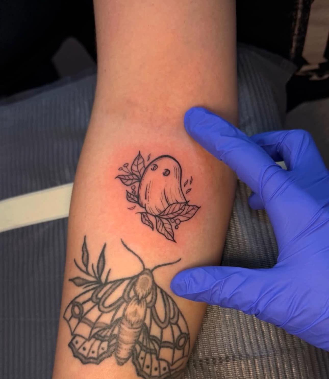 Blue-gloved hand pointing to a small bird tattoo above a larger moth tattoo on a forearm