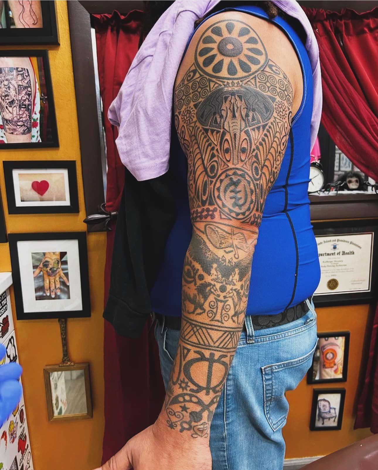 Person with full-arm tattoos wearing a blue top and jeans, standing before a wall of framed artwork.