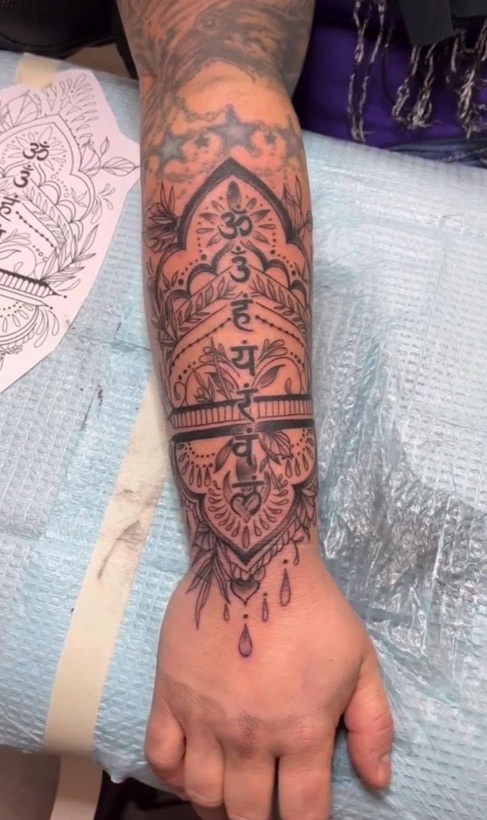Arm with a rectangular black-outline tattoo showing a mountain and stag, beside a bed and colorful wall art.