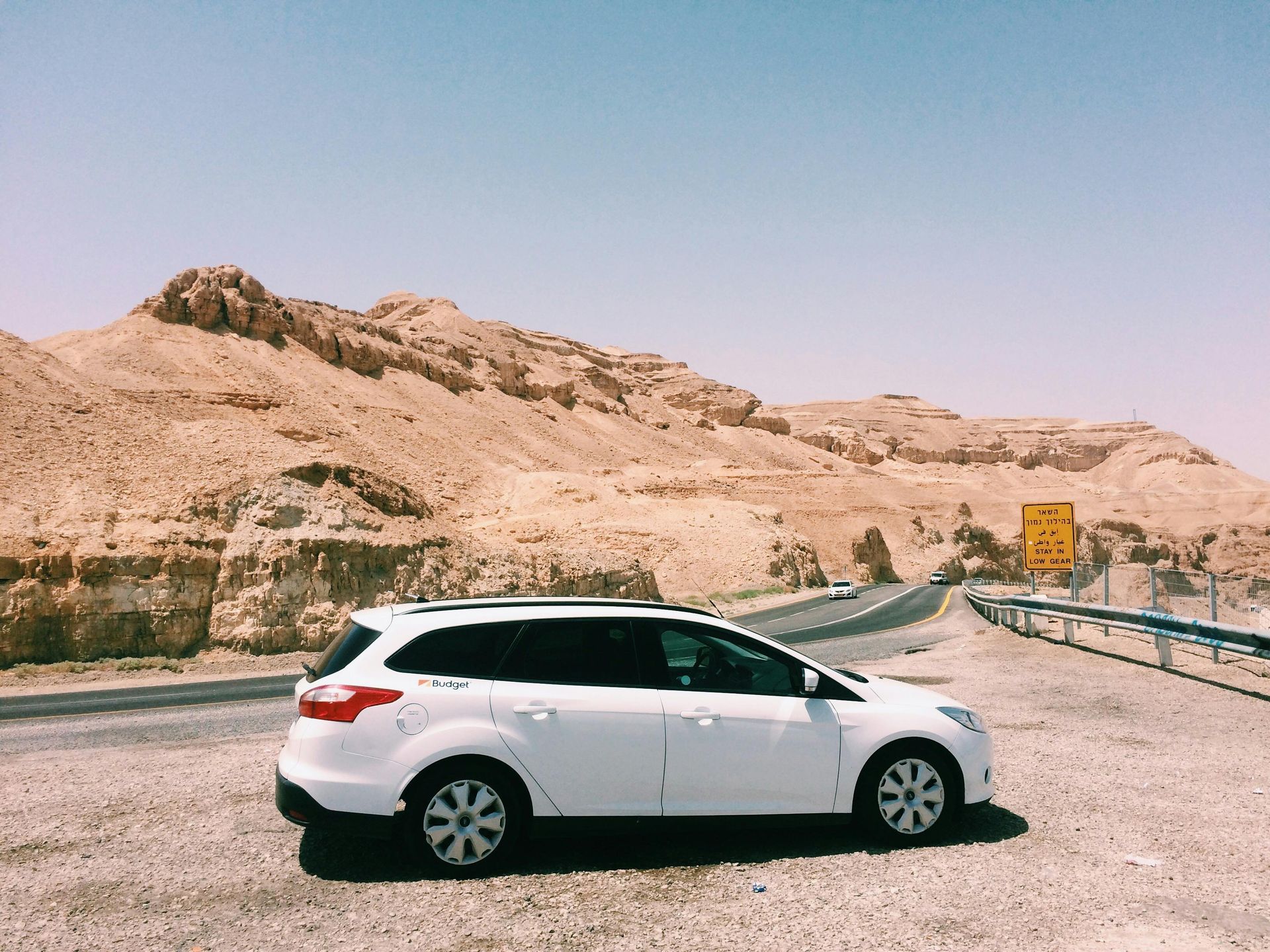 White car parked on side of desert road, tan mountains in the background, blue sky.
