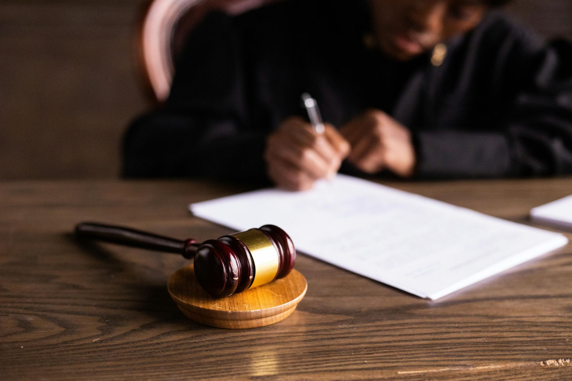 Gavel on wooden block in foreground, judge writing on document in background.