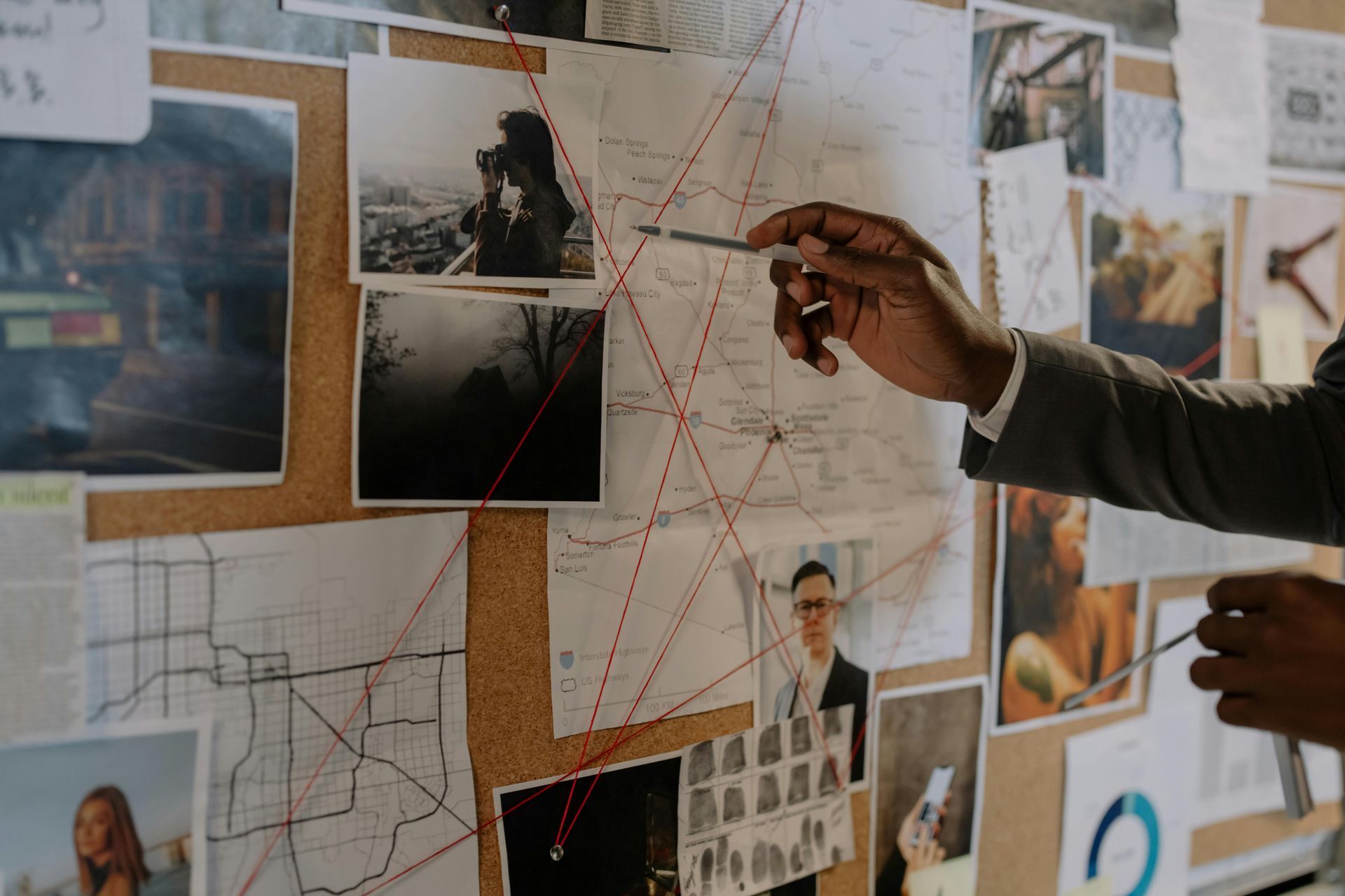 Person points at connected photos, maps on a corkboard, suggesting an investigation.
