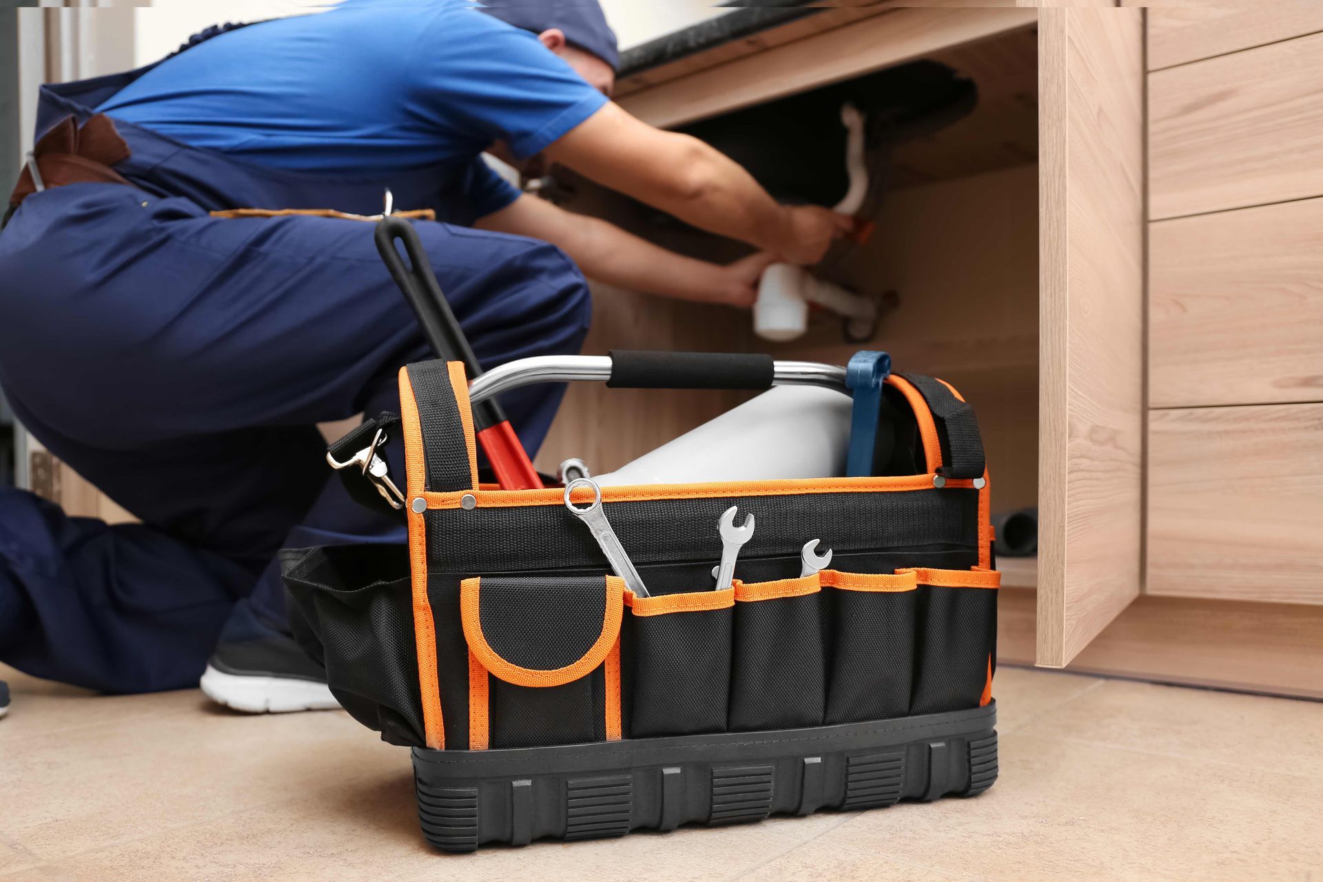 Technician in blue uniform working under a cabinet beside a black and orange tool bag