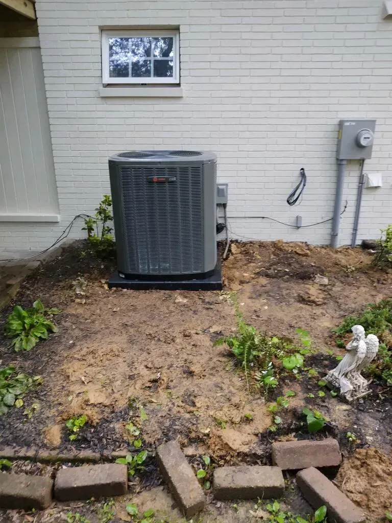 Outdoor HVAC unit beside a house wall over bare dirt, with a small window and utility meter nearby.
