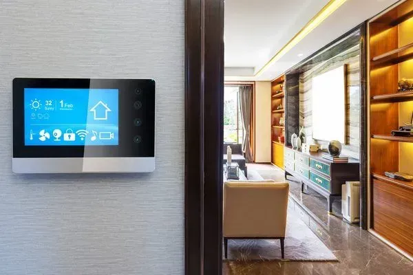 Wall-mounted touchscreen control panel beside a modern hotel room interior with sofa and window seating.