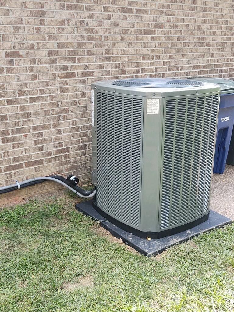 Outdoor air conditioning unit beside a brick wall on a grassy lawn, with a hose along the ground.