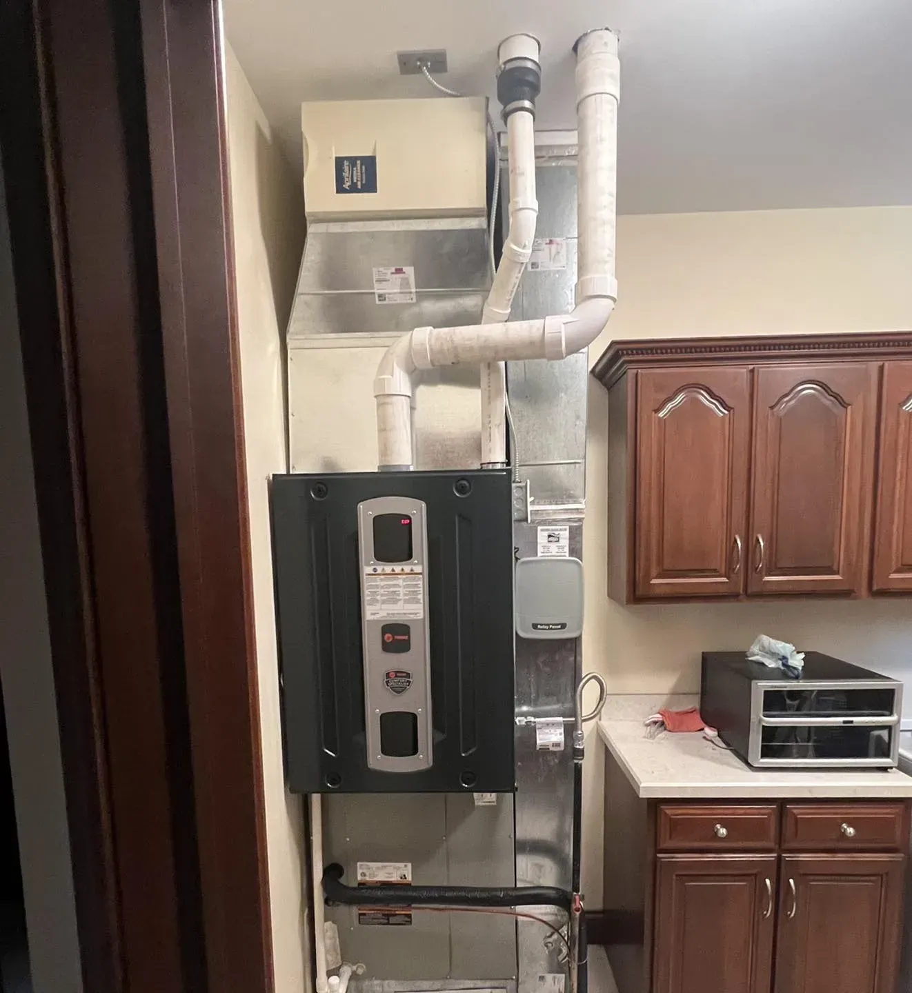 Indoor utility room with a wall-mounted boiler and white pipes beside wooden kitchen cabinets.