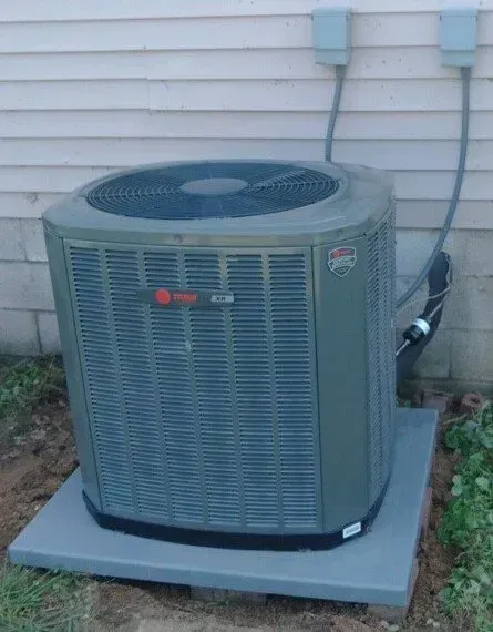 Outdoor central air conditioner unit beside a house wall on a concrete pad.