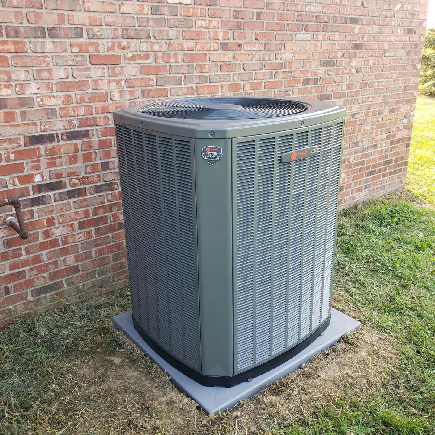 Outdoor air conditioning unit beside a brick wall on a concrete pad with grass around it