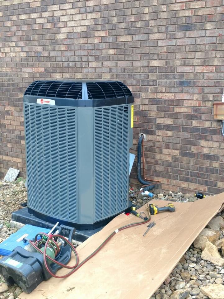 Outdoor HVAC unit on plywood beside a brick wall, with tools and hoses nearby.
