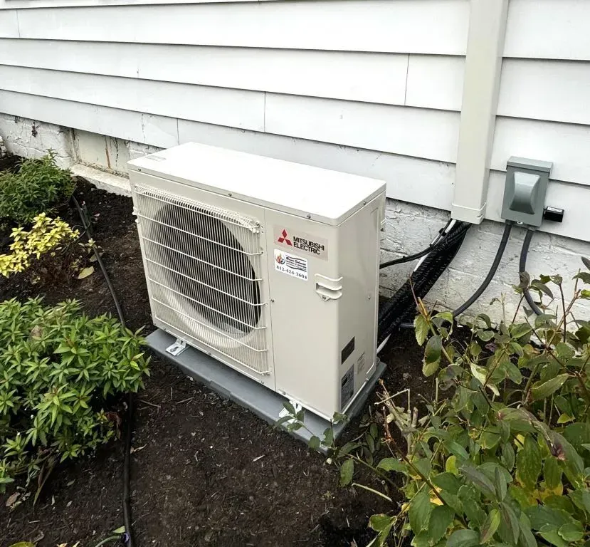 Outdoor air-conditioning unit beside a white house, with hoses connected near the wall.