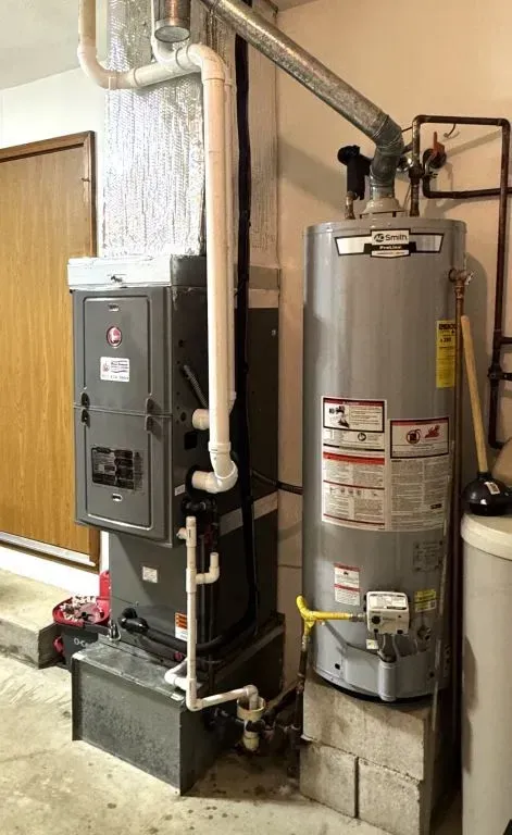 Two gray water heaters and a gray electrical panel in a utility room with exposed pipes and cinder blocks.