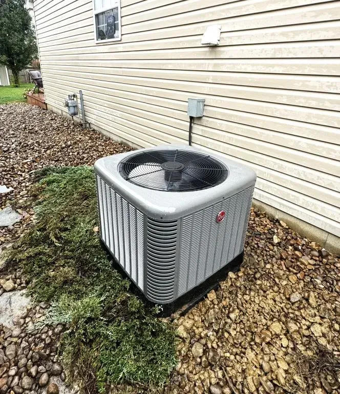 Outdoor air conditioning unit beside a beige house, sitting on gravel and grass.