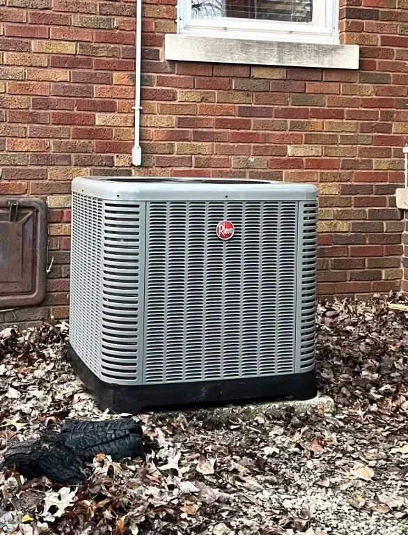 Outdoor central air conditioning unit beside a brick house wall, surrounded by fallen leaves.