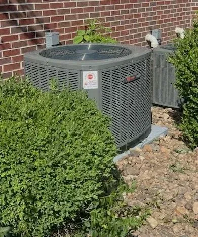 Outdoor HVAC unit beside a brick wall, partially hidden by bushes and rocks.