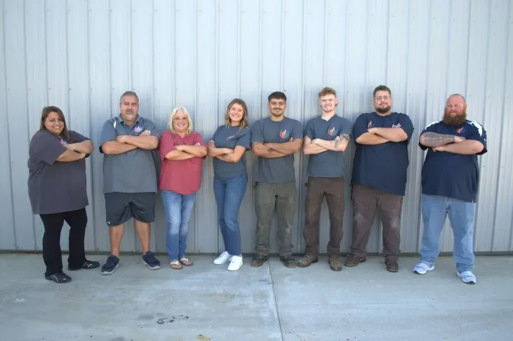 Seven people standing shoulder-to-shoulder with arms crossed in front of a gray metal wall.