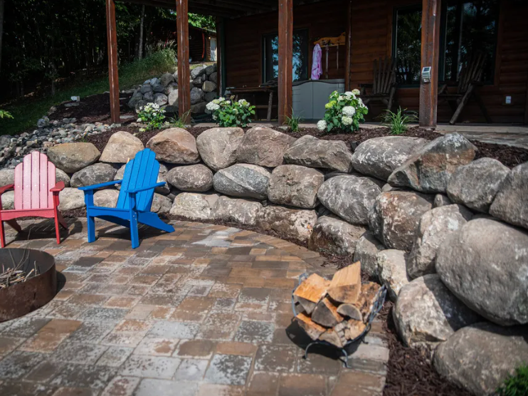 Stone patio with red and blue chairs, stacked rocks, and a small fire pit in front of a wooden cabin.