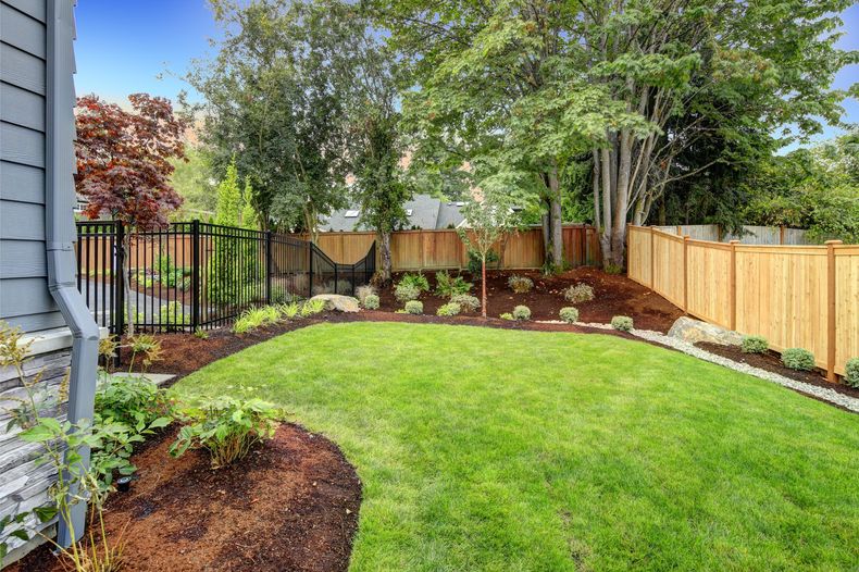 Landscaped backyard with green lawn, flower beds, wooden fence, and trees under bright daylight