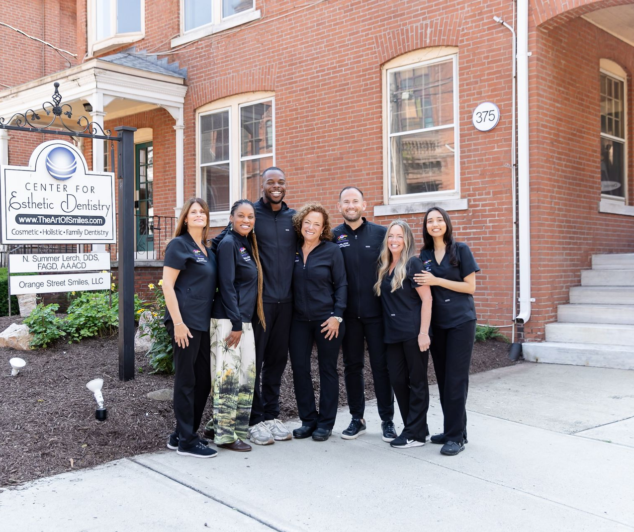 Six people in black attire stand outside a brick building beside a church sign.
