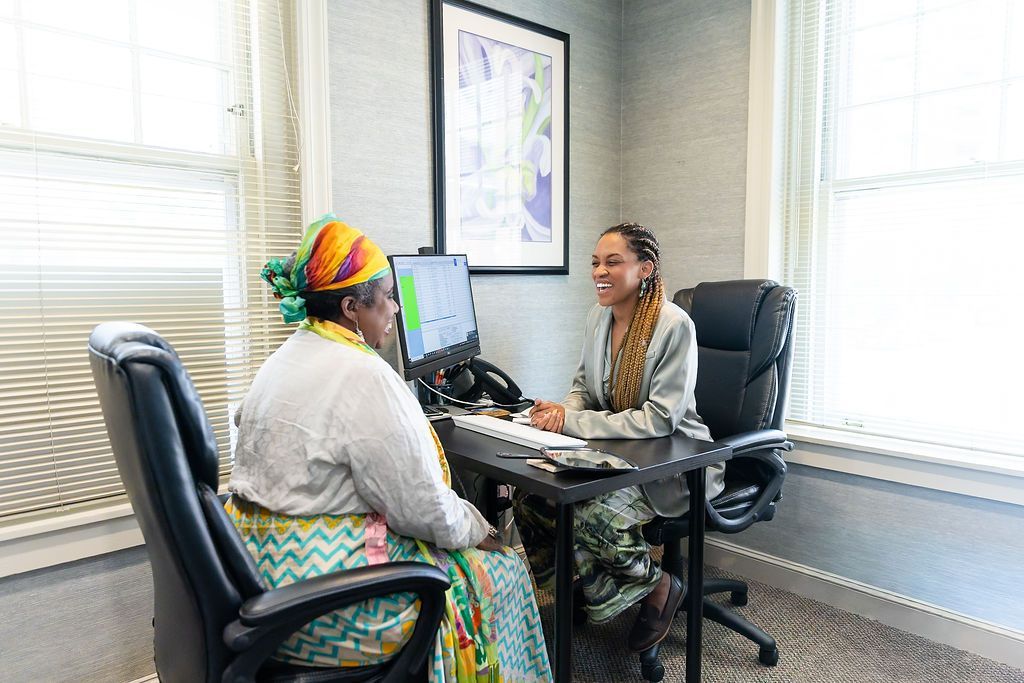 Two people talking across a desk in an office, with a computer between them and bright windows behind.