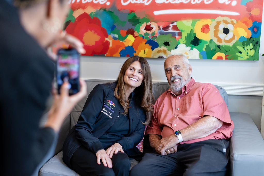Two people sit on a couch smiling while someone in the foreground takes their photo.