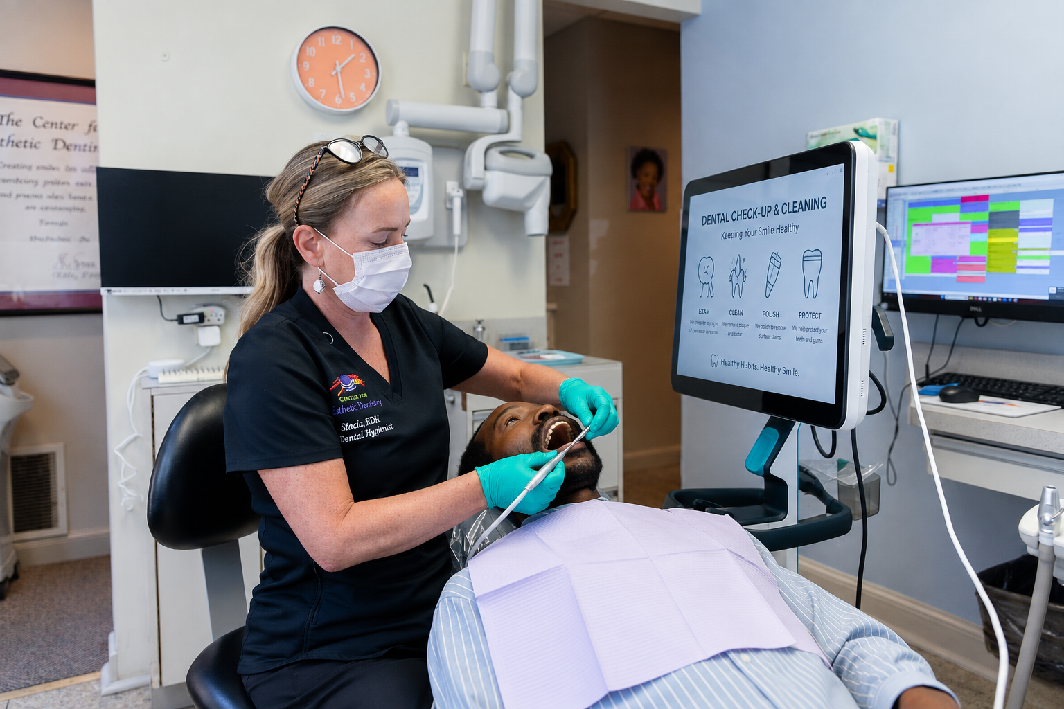 Dentist examining a patient in a chair beside a monitor showing dental images