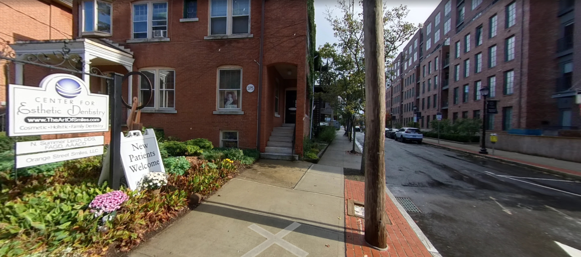 Brick apartment building entrance with sign, flowers, and sidewalk along a quiet street